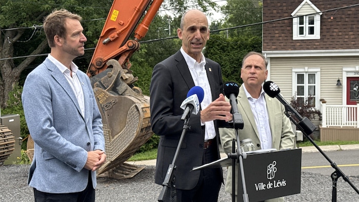 Olivier Biron, Steven Blaney et Jean Demers devant un lutrin en conférence de presse, derrière eux une pelle mécanique.