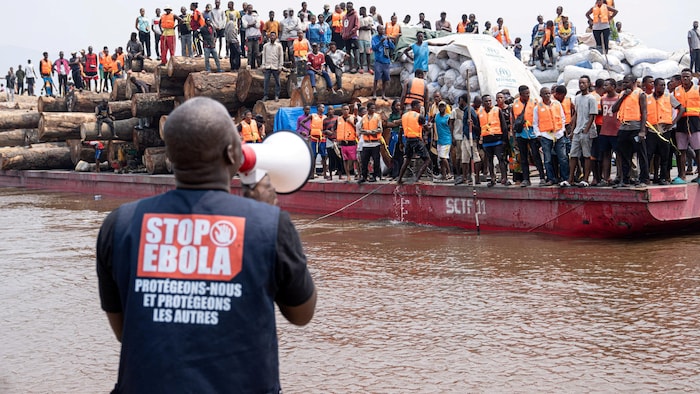 Un homme de dos, portant un chandail avec les mots «Stop Ebola, protégeons-nous et protégeons les autres», parle dans un mégaphone à plus d'une centaine de personnes sur un navire rempli de passagers.
