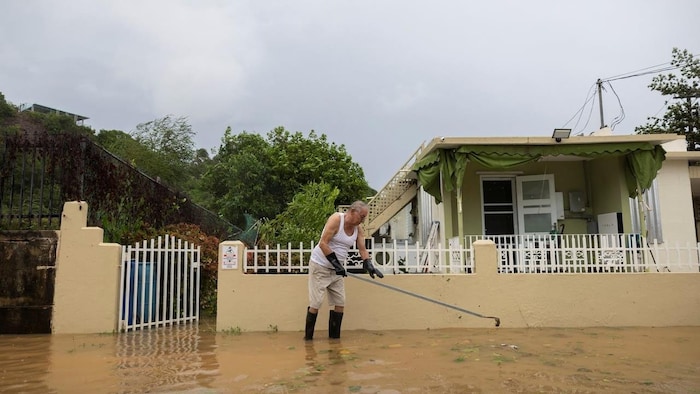 Um homem usando botas de chuva, atravessando vários centímetros de água, usa uma tocha para desentupir um cano na frente da casa.