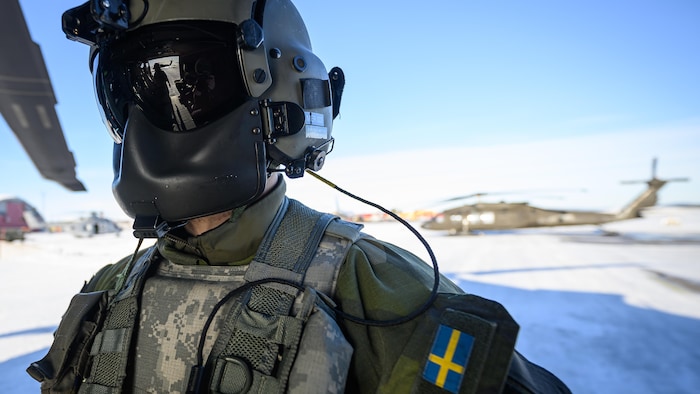 Un soldat masqué près d'un hélicoptère. Son uniforme porte un drapeau suédois.