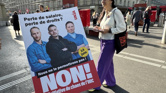 Une femme debout à côté d'une pancarte où l'on peut lire « Perte de salaire, perte de droits? Non! ».