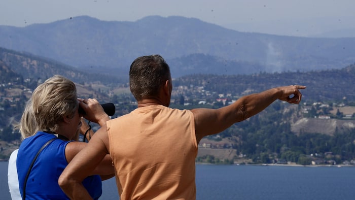 Des personnes rassemblées à un belvédère aménagé surplombant le lac Okanagan. La rive opposée et les collines entourant Summerland se découpent à travers un ciel voilé par la fumée de feu de forêt.