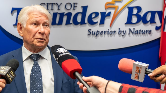Ken Boshcoff en train de répondre à des questions de journalistes avec trois micros devant lui.