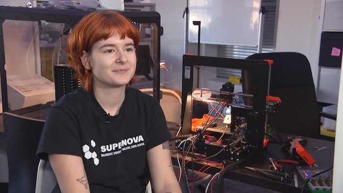 Une femme avec un t-shirt noir de SuperNOVA est assise à côté d'une table couverte de fils et d'appareils électroniques.
