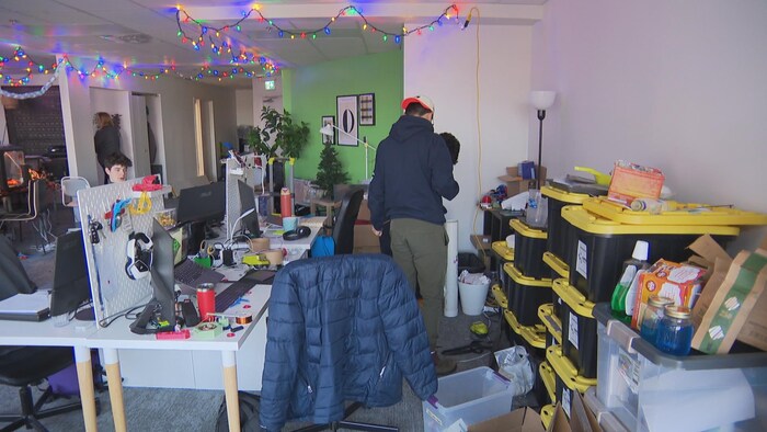 Un bureau rempli d'objets divers qui jonchent les tables et le sol. Il y a des contenants de rangement en plastique noir et jaune et des lumières de Noël allumées au plafond.