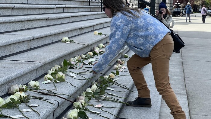 Une dame dépose une roseate  blanche sur des escaliers. Plusieurs roses sont déjà déposées, avec une étiquette determination    un nom sur chaque tige. 