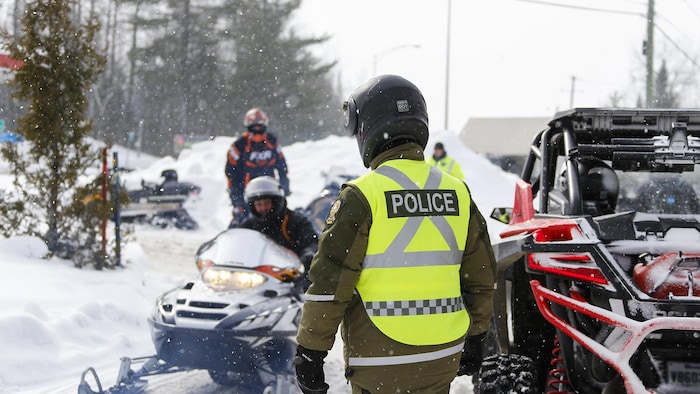 Un policier avec un casque s'approche de plusieurs motoneigistes, en hiver.