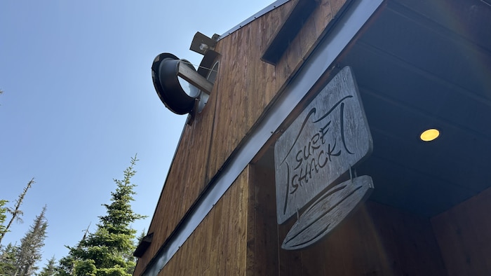 Une enseigne en bois suspendue à une structure sur laquelle est inscrit « Surf Shack » avec le dessin d'une planche de surf en dessous, se détachant sur un ciel.