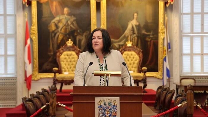 Susan Corkum-Greek debout au lutrin devant les somptueux tableaux que l'on retrouve dans l'édifice de l'Assemblée législative.