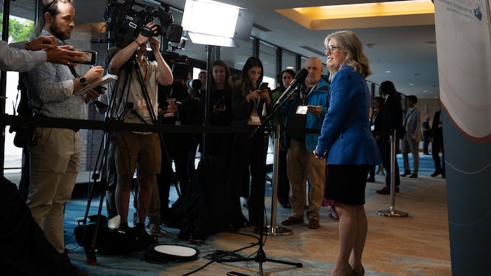 Susan Holt debout devant un micro sur pied. Elle fait face à un mur de journalistes et à un caméraman.