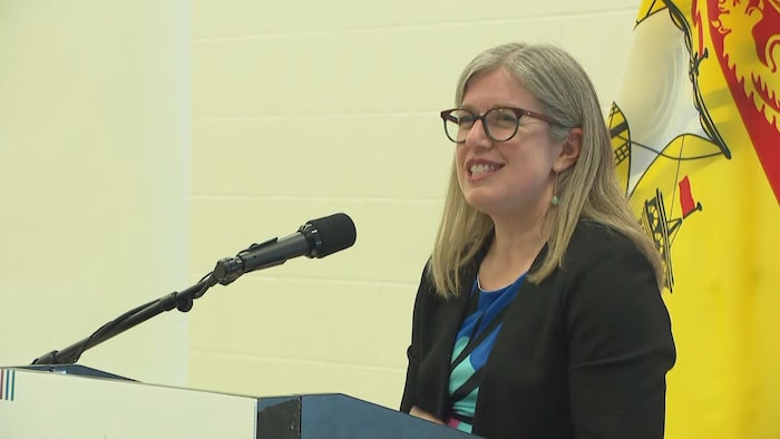 La première ministre Holt derrière un micro, devant un drapeau du Nouveau-Brunswick. 