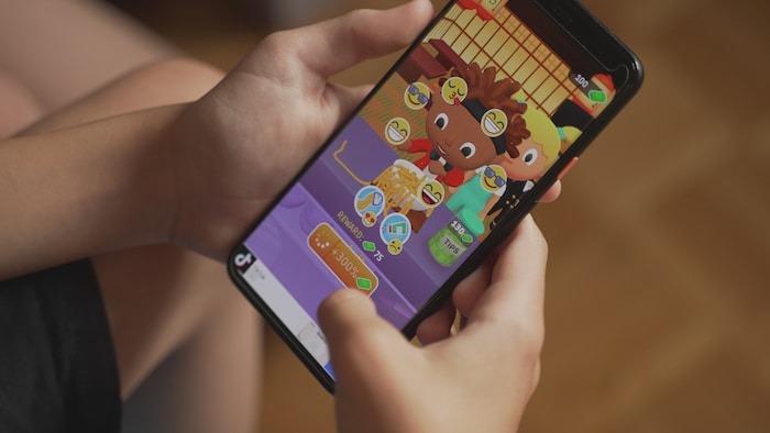 Des mains d'enfant tiennent un téléphone intelligent affichant un jeu mobile coloré, avec des émojis.