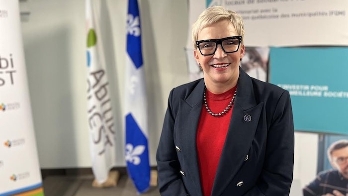 Suzanne Blais sourit à la caméra en posant devant des drapeaux du Québec et de la MRC d'Abitibi-Ouest.