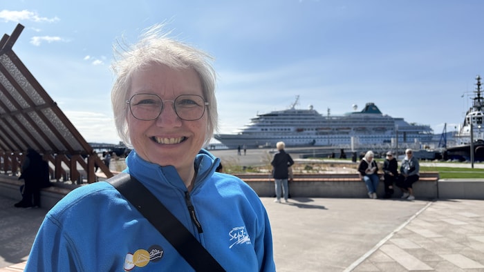 Un portrait à mi-corps d'une femme souriante portant des lunettes et une veste polaire debout à l'extérieur. Derrière elle, on voit le grand paquebot MS Amera à quai, ainsi que des passagers assis sur des bancs au loin.