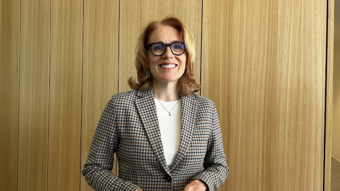Une femme souriante aux cheveux roux, portant des lunettes de vue et un veston à carreaux sur un haut blanc.