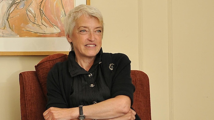 Une femme est assise sur un petit divan. Elle a les bras croisés et elle sourit.