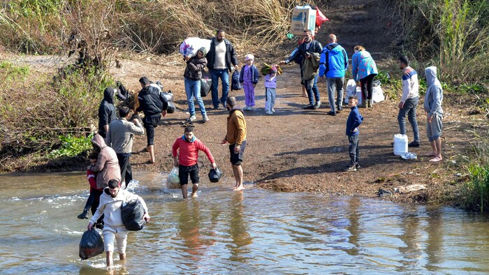 Des familles portant des bagages traversent une rivière.