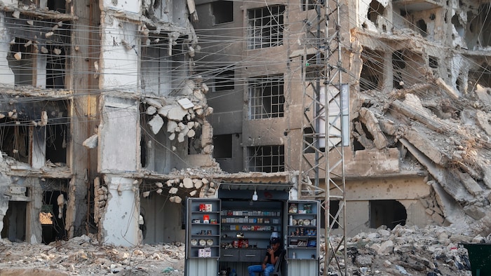 Un homme attend des clients devant un petit étalage placé à côté de grands bâtiments à moitié détruits.