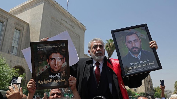 Au milieu d'une foule devant un palais de justice, un homme en complet brandit deux photos encadrées.