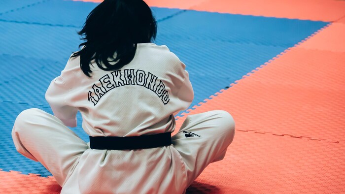 Une jeune fille vue de dos portant une tenue d'entraînement avec l'inscription « taekwondo » sur son dos est assise sur un matelas coussiné.