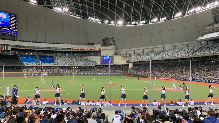 Une vue du champ extérieur et des écrans géants du Taipei Dome pendant un match de baseball, avec des meneuses de claques à l'avant-plan.