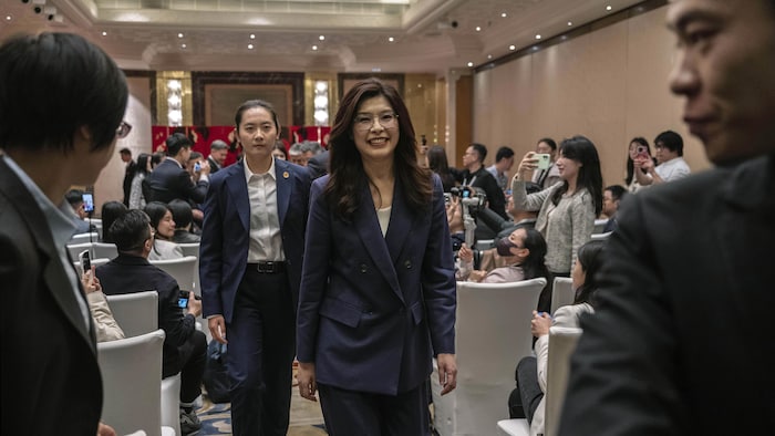 Une femme en costume bleu marche dans une salle, suivie d'un homme, et est prise en photo par des dizaines de personnes.