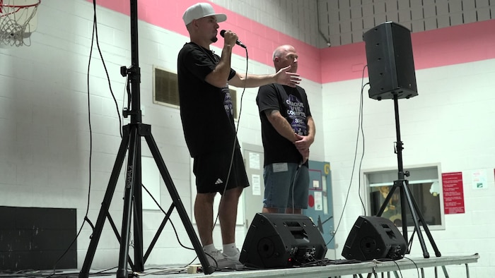 Les rappeurs Simon Valiquette et Frédéric Auger du groupe Taktika debout sur une petite scène dans un gymnase, entourés de haut-parleurs. Frédéric parle au micro.
