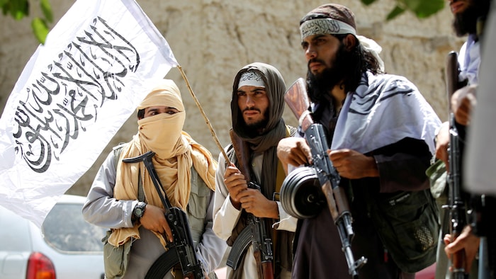 Quatre talibans armés de mitraillettes brandissent un drapeau dans une rue.