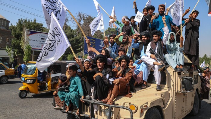 Une vingtaine d'hommes et de jeunes garçons, assis sur un véhicule blindé avec un homme armé, brandissent des drapeaux de l'Émirat islamique d'Afghanistan pendant un défilé.