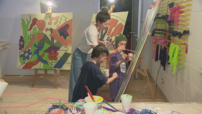 Une femme et deux enfants peignent ensemble sur de grands chevalets dans un atelier d'art. Des pots de peinture colorée et des pinceaux sont disposés au premier plan, tandis que des œuvres colorées et textiles décorent le mur.