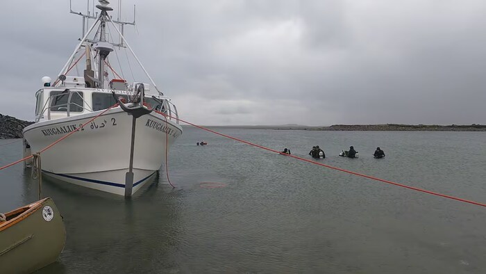 Un bateau de pêche est amarré dans des eaux calmes sous un ciel gris, tandis qu’un groupe de plongeurs en combinaison noire évolue à proximité, relié au navire par une longue corde rouge.
