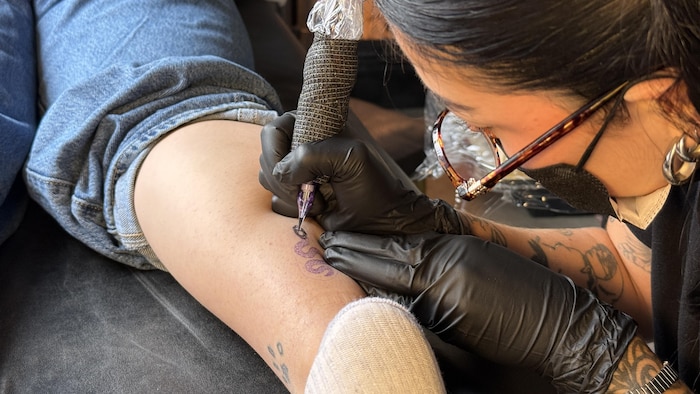 Marissa Couturier trace les lignes d'un tatouage avec une machine à tatouer sur un mollet.