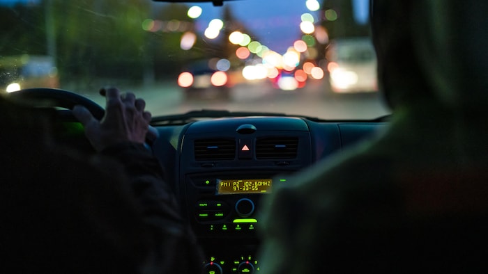 Photo de l'intérieur d'un taxi la nuit. On voit la main du chauffeur sur le volant et la silhouette d'un passager assis à l'arrière.