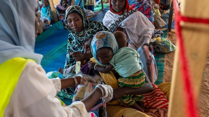 Une femme mesure le bras d'une femme qui tient son bébé, dans une file de femmes assises sur une couverture par terre.