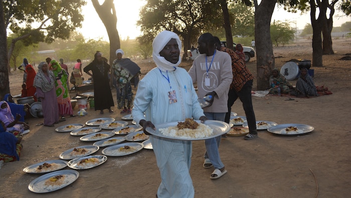 Un homme porte une assiette de nourriture, tandis que plusieurs autres sont posées par terre derrière lui.