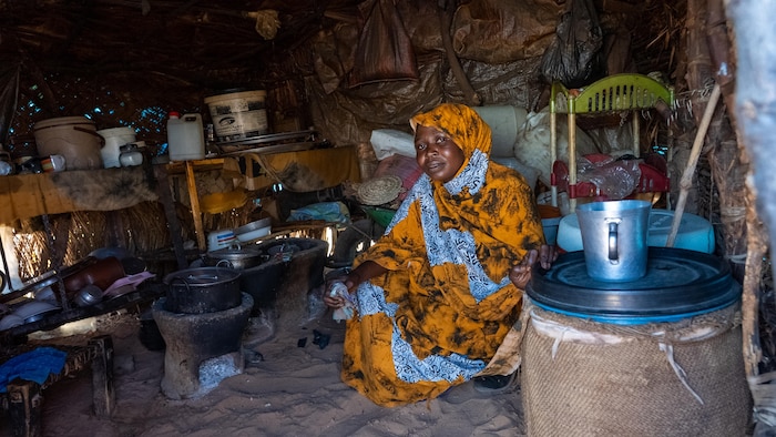Zouhour Mahamad, en tenue traditionnelle, est assise sur le sable à côté de ses chaudrons dans un abri.