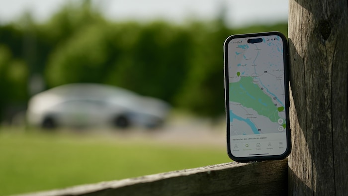Un téléphone sur lequel s'affiche la carte des stations de Communauto dans la MRC des Collines-de-l'Outaouais est posé contre une clôture devant une voiture Communauto à l'arrière-plan.