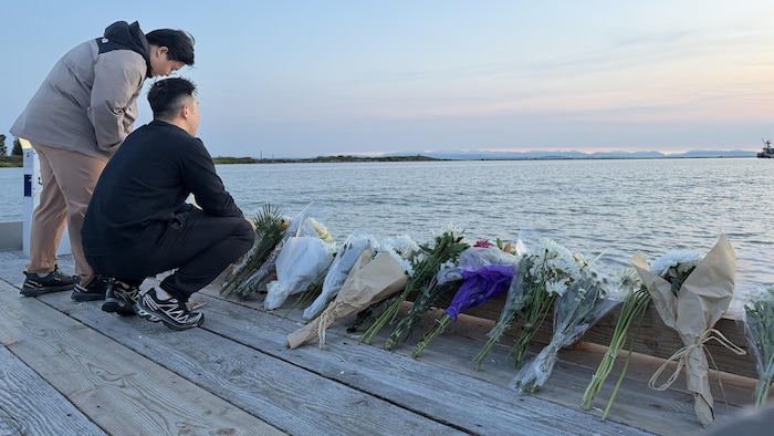 Des bouquets de fleurs sont posés sur un quai, et deux personnes regardent la mer.