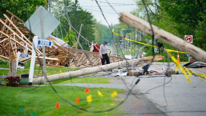 Des poteaux électriques et des structures en bois jonchent le sol après une tempête. Une personne est au milieu pour nettoyer.