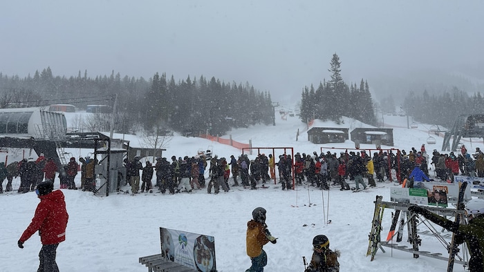 Une longue file de skieurs à l'entrée du remonte-pente d'une station de ski. Le ciel est couvert et de la neige tombe.
