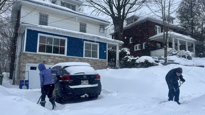 La neige tombe et deux personnes pellettent leur entrée.