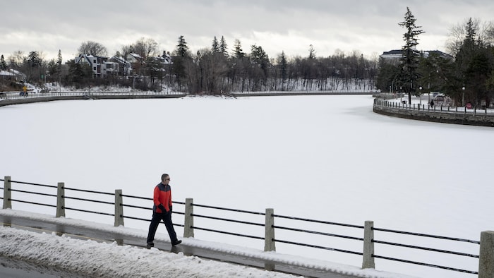 Un homme marche le long d'un canal enneigé qui est normalement une patinoire l'hiver mais qui est fermé faute de froid.