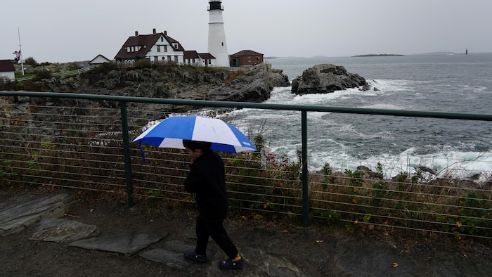 A small boy with an umbrella is in front of the ocean and the fence in front of a lighthouse.