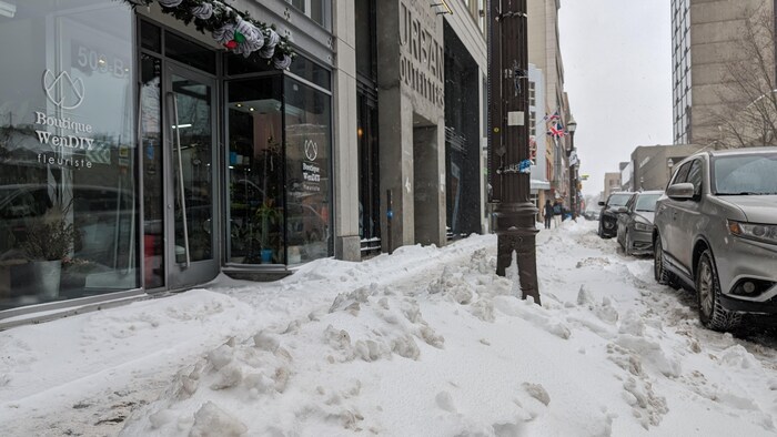 Un épais amas de neige sépare le trottoir de la chaussée dans une rue du centre-ville où des voitures sont stationnées. 