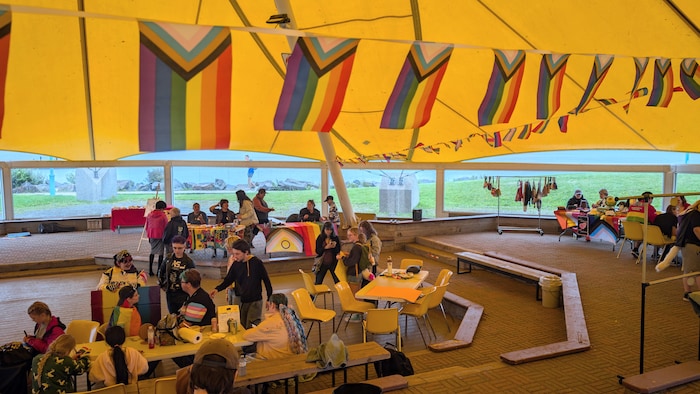 Des personnes sont installées autour de tables sous la tente jaune décorée de guirlandes de drapeaux arc-en-ciel.