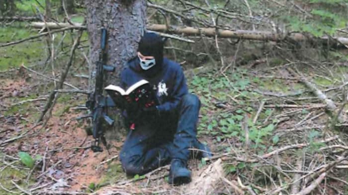 Un membre masqué de la Division Atomwaffen en train de lire contre un arbre avec un fusil d’assaut à ses côtés.