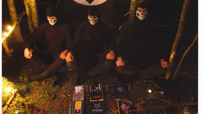 Trois hommes masqués et vêtus de noir assis dans une forêt devant un drapeau nazi.