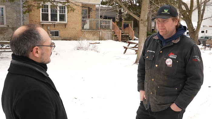 Le pompier Shane Kirby porte un manteau d'hiver et une casquette. Il discute avec le journaliste Christian Noël dans une cour enneigée.