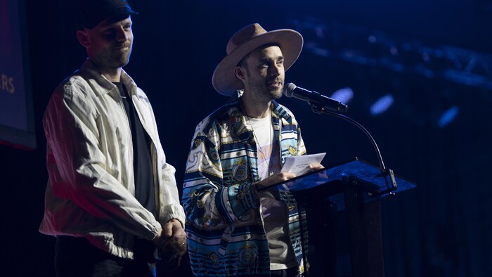 Deux hommes sur scène devant un micro. L'un d'eux prononce quelques mots en acceptant un prix.