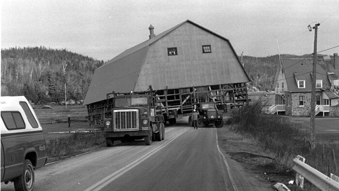 Photo d'archives en noir et blanc. Deux camions tirent une grange sur une route.